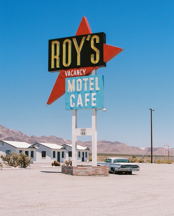 A vintage sign for Roy's Motel and Cafe stands in a desert landscape with a classic car parked nearby.