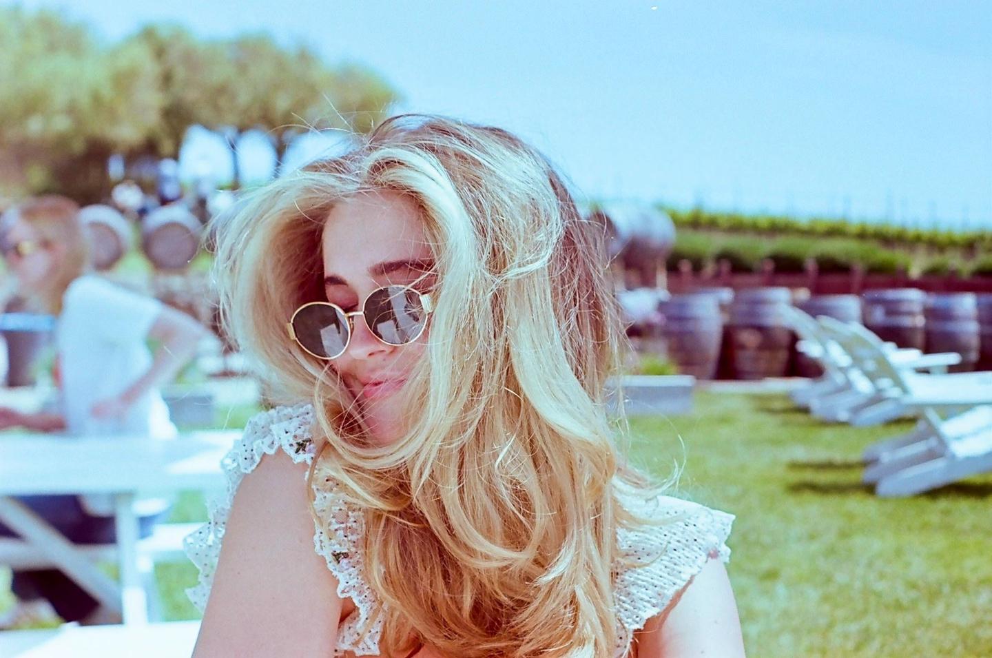 A woman with long blonde hair and sunglasses smiles while sitting outdoors with wine barrels in the background.