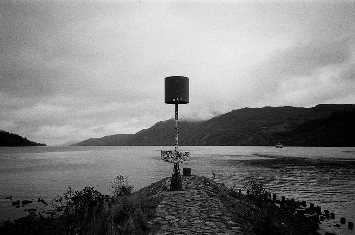 A black and white landscape shows a lake with mountains in the background and a cylindrical marker in the foreground.