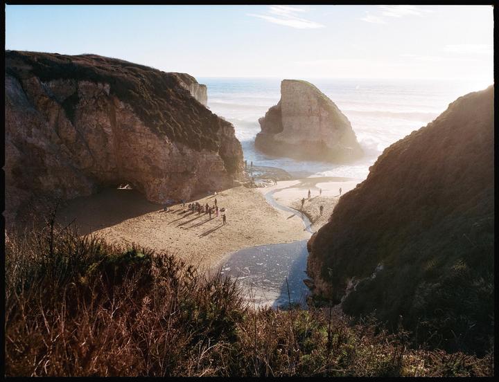 A group of people are gathered on a sandy beach between two large cliffs, with the ocean and a sea stack in the background.