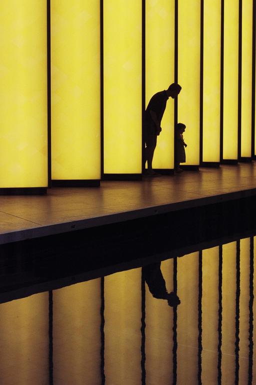 A man and a child are silhouetted against a wall of yellow light panels, with their reflections visible in a dark surface below.