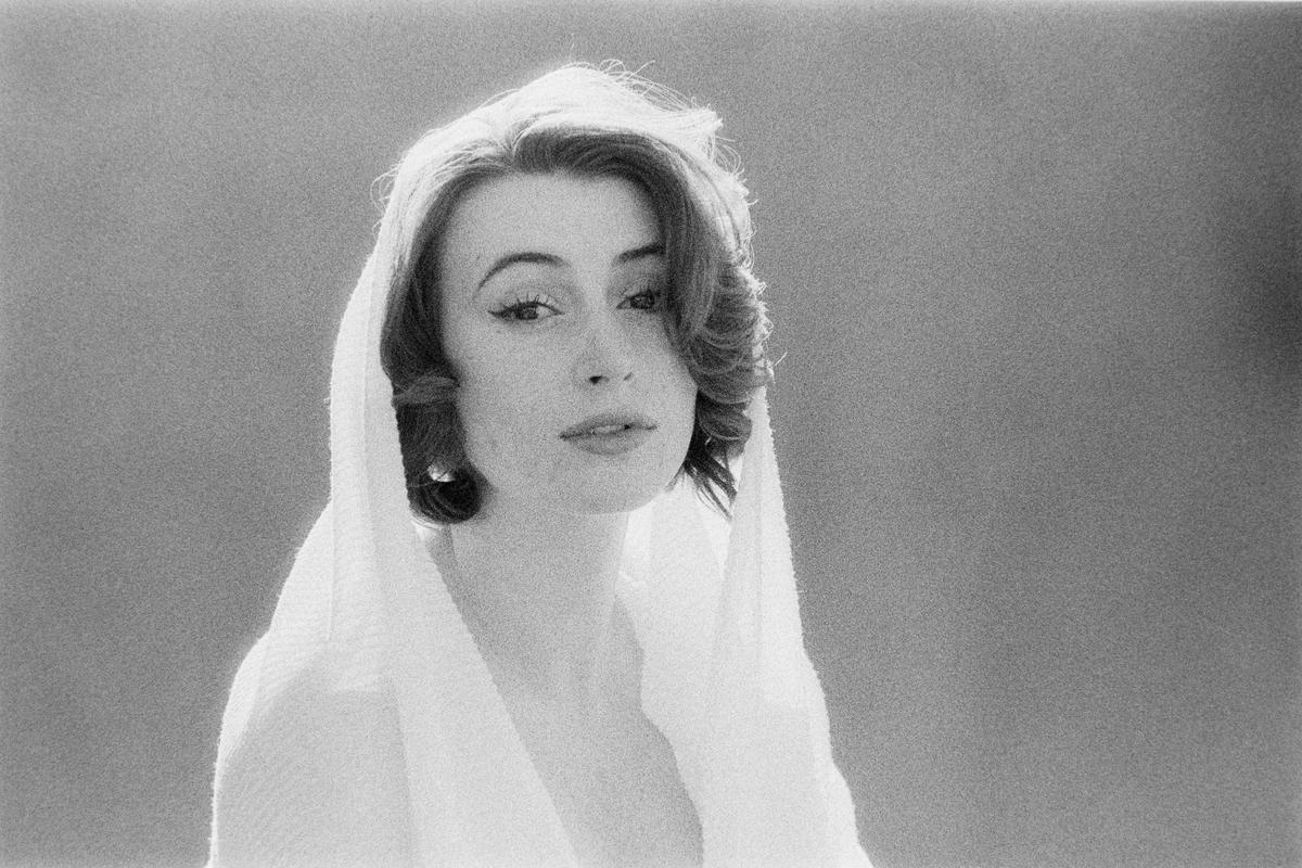 A woman with dark, curly hair and winged eyeliner looks directly at the viewer while wearing a white, textured hood.