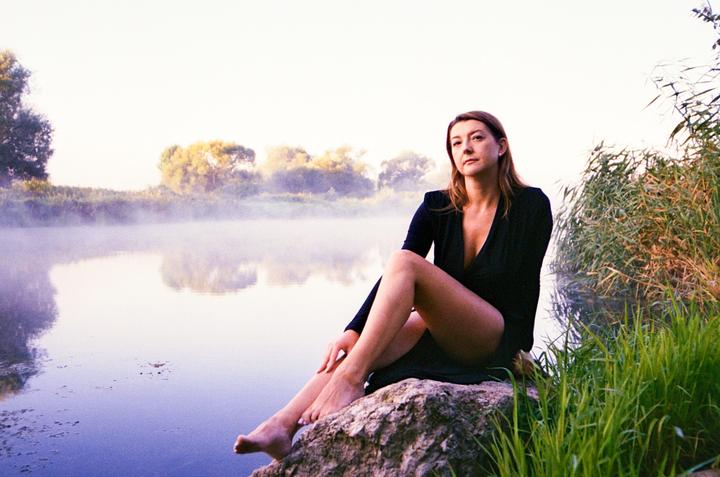 A woman in a black dress sits on a rock by a misty lake with tall grass and trees in the background.
