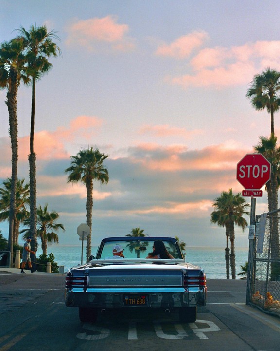 A convertible car with two people inside is stopped at an intersection overlooking the ocean at sunset, with palm trees and a stop sign visible.