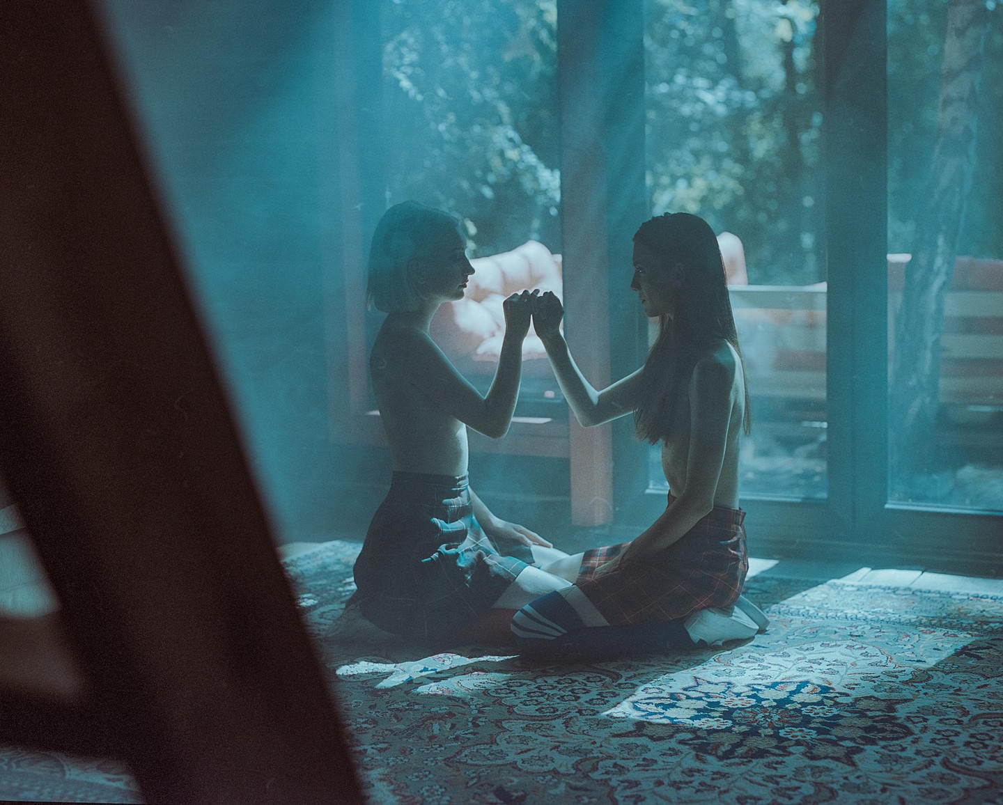 Two women sit on a patterned rug, holding hands and facing each other in a room with large windows.
