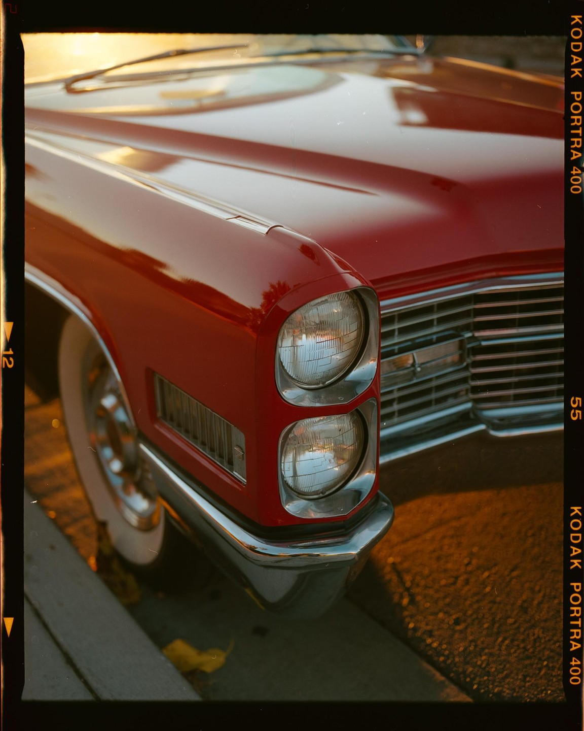 The front of a red vintage car is parked on a street during golden hour.