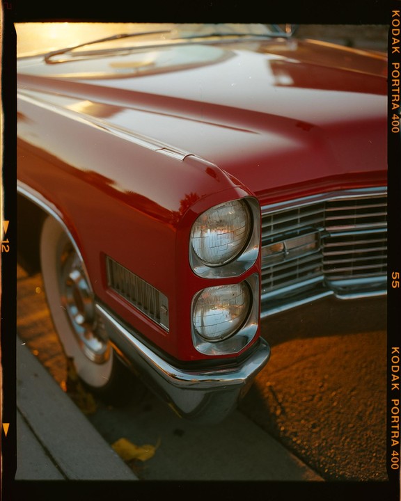 The front of a red vintage car is parked on a street during golden hour.