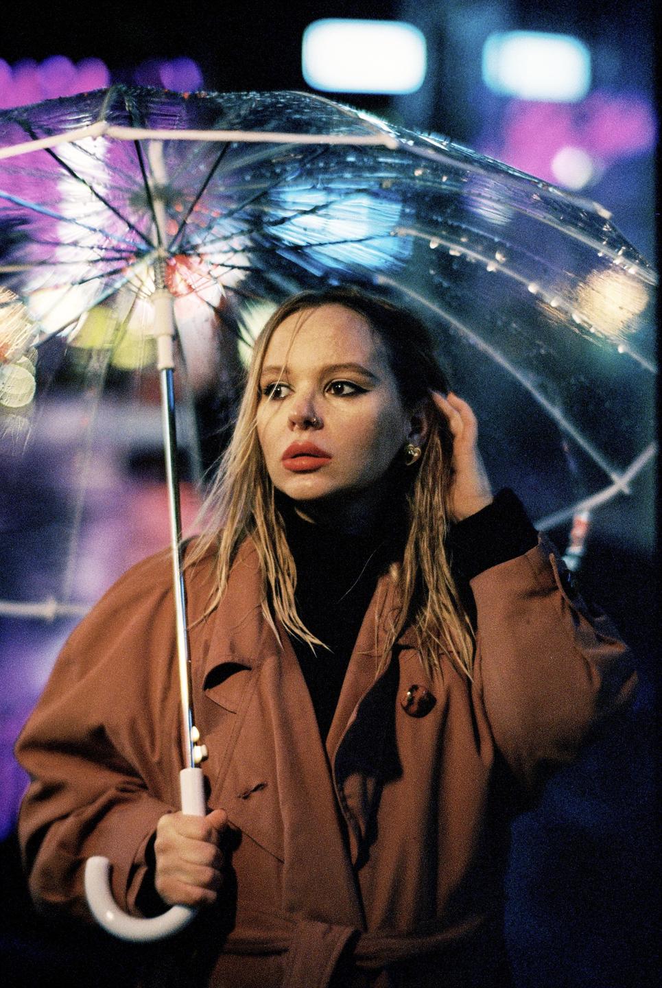 A woman in a trench coat holds a clear umbrella at night.