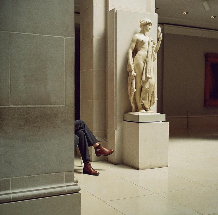 A person's legs and feet are visible behind a stone column, next to a classical statue of a woman holding a mirror.