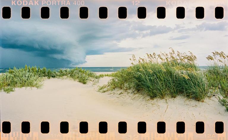 A shelf cloud looms over a sandy beach with sea oats and ocean waves.