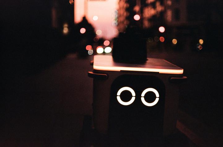 A delivery robot with glowing circular lights on its front is visible at night with blurred city lights in the background.