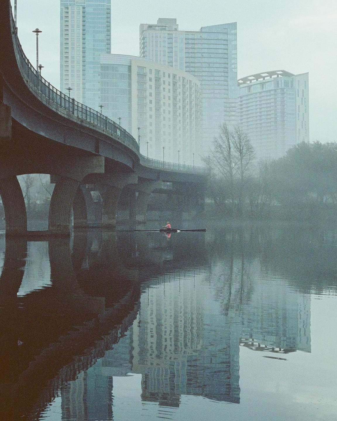 A person in a scull rows on a river with a bridge and tall buildings in the background.