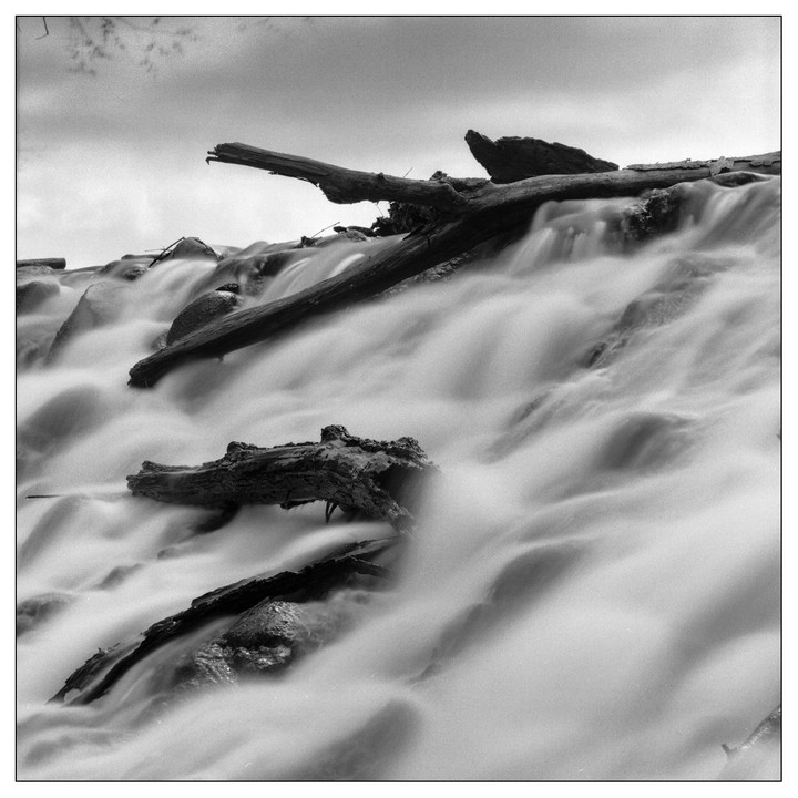 A long exposure captures the smooth flow of water over rocks and logs in a stream.