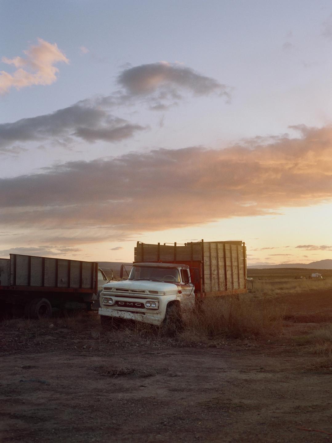 An old white GMC truck with a wooden bed is parked in a field at sunset.
