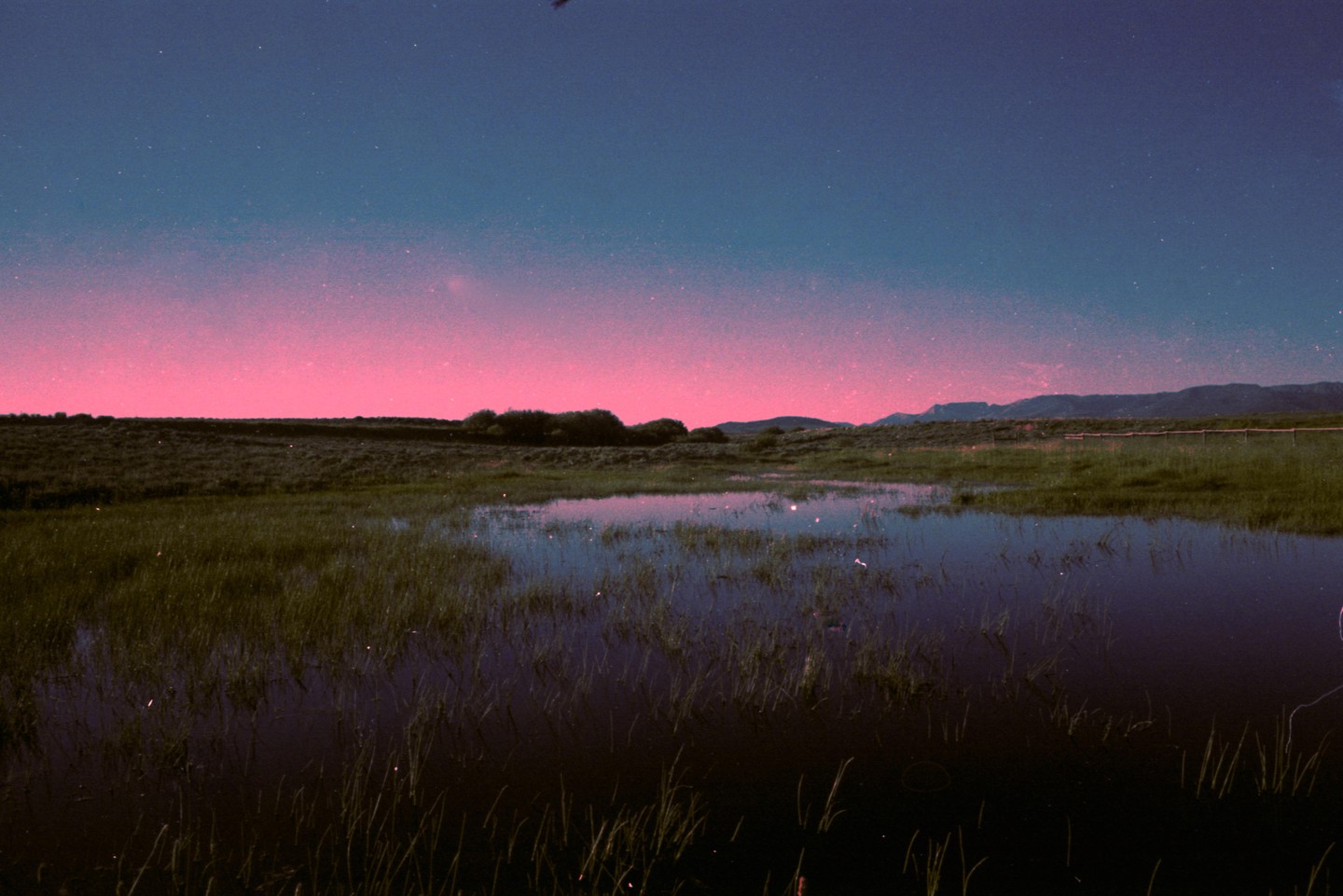 A pink and blue sky with stars is reflected in a body of water surrounded by grass and distant mountains.