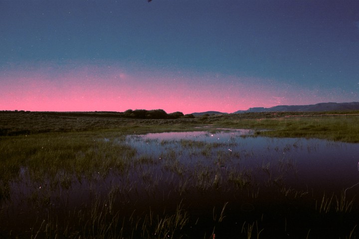 A pink and blue sky with stars is reflected in a body of water surrounded by grass and distant mountains.