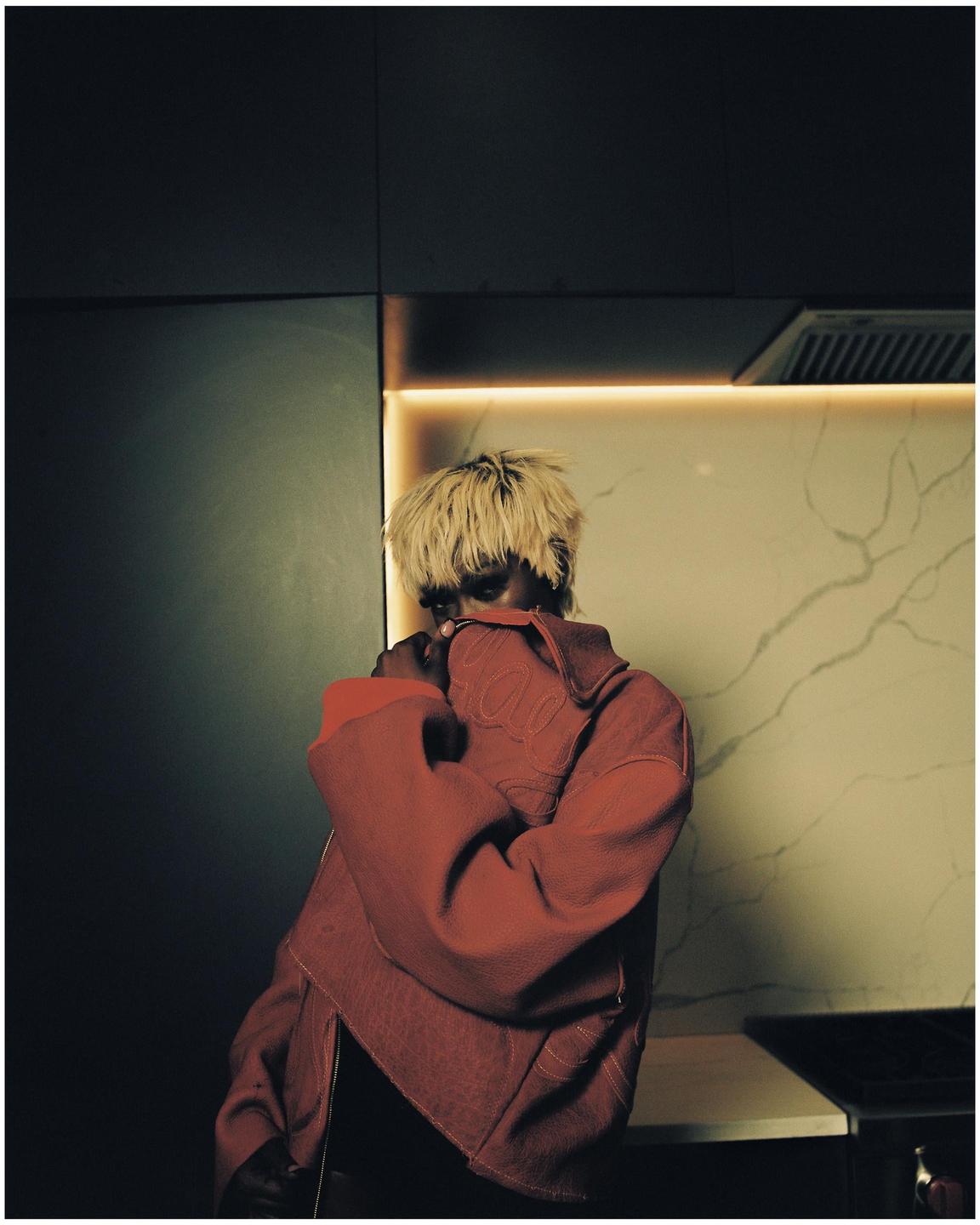A person with bleached hair and a red jacket stands in a kitchen with illuminated countertops.