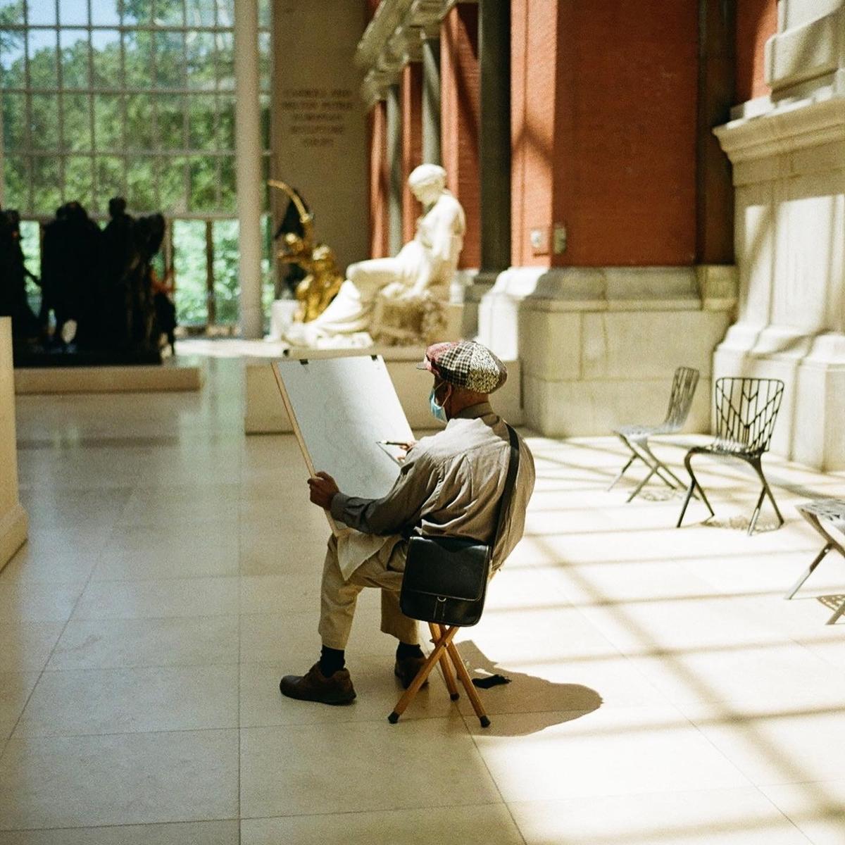 A man wearing a face mask and a plaid cap sits on a stool and draws on an easel in a museum.