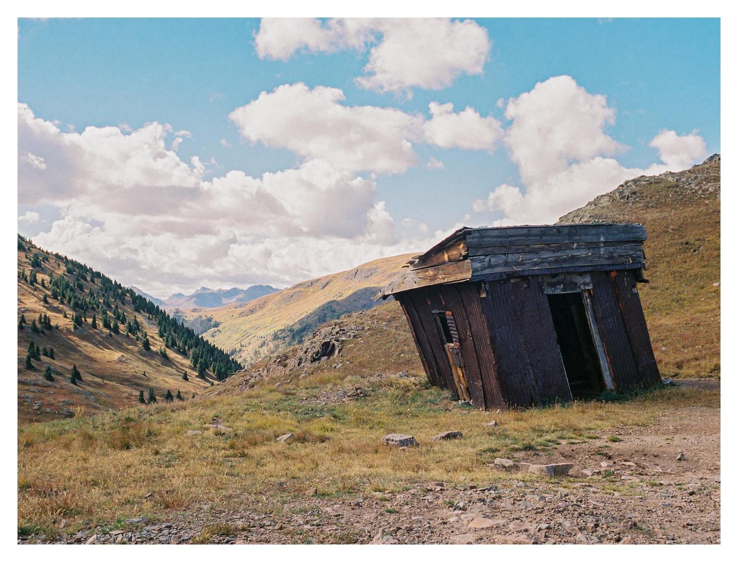 An old, leaning corrugated metal shack sits on a grassy hillside with mountains and a valley in the background under a cloudy sky.