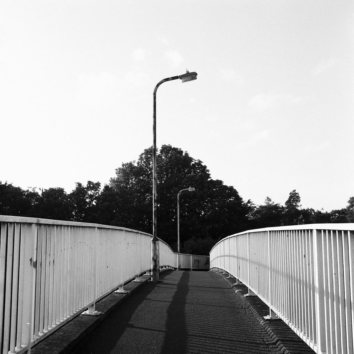 A pedestrian bridge with white railings curves towards a dark tree line under a bright sky.