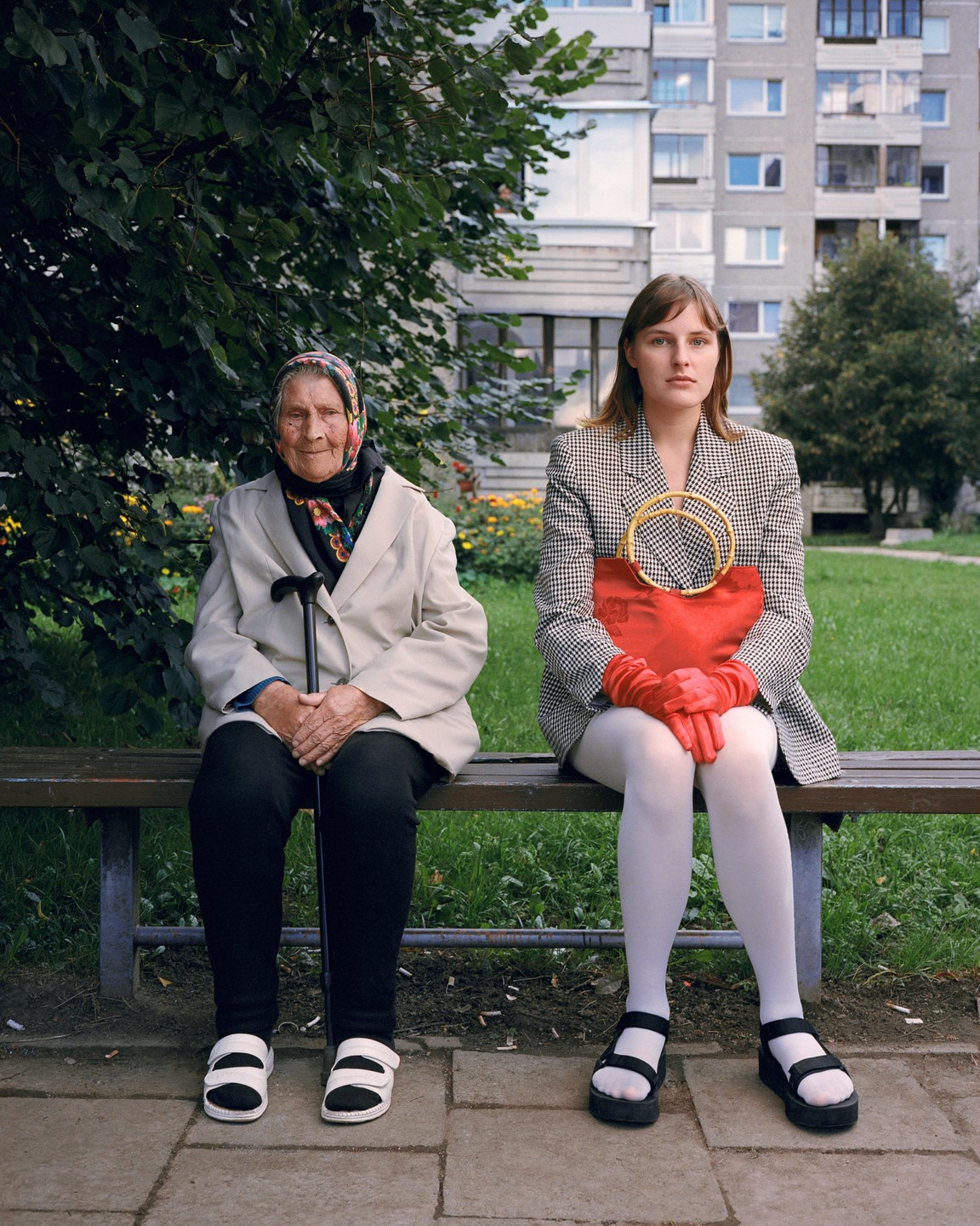 An older woman and a young woman sit side-by-side on a park bench in front of an apartment building.