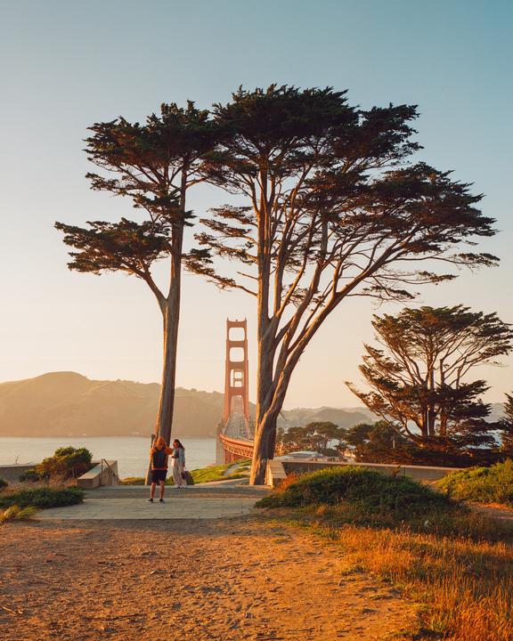 Two people stand on a path overlooking the Golden Gate Bridge at golden hour, framed by two large trees.