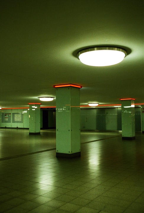 An empty subway station features green pillars with orange tops and circular ceiling lights.