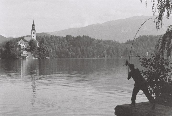 A man fishes in a lake with a church on an island and mountains in the background.