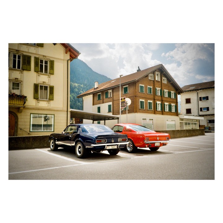 Two classic cars are parked in a lot in front of buildings with mountains in the background.