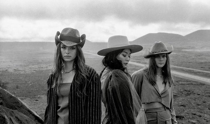 Three women wearing hats stand in a barren landscape with mountains in the distance.