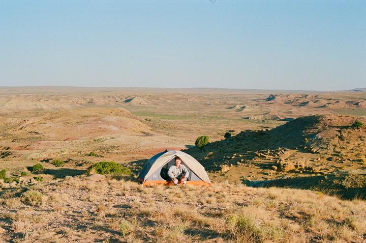 A man sits inside a tent pitched on a dry, grassy hill overlooking a vast desert landscape.