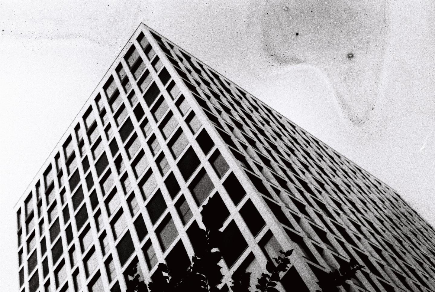 A tall building with a grid pattern of windows is seen from a low angle against a bright sky.