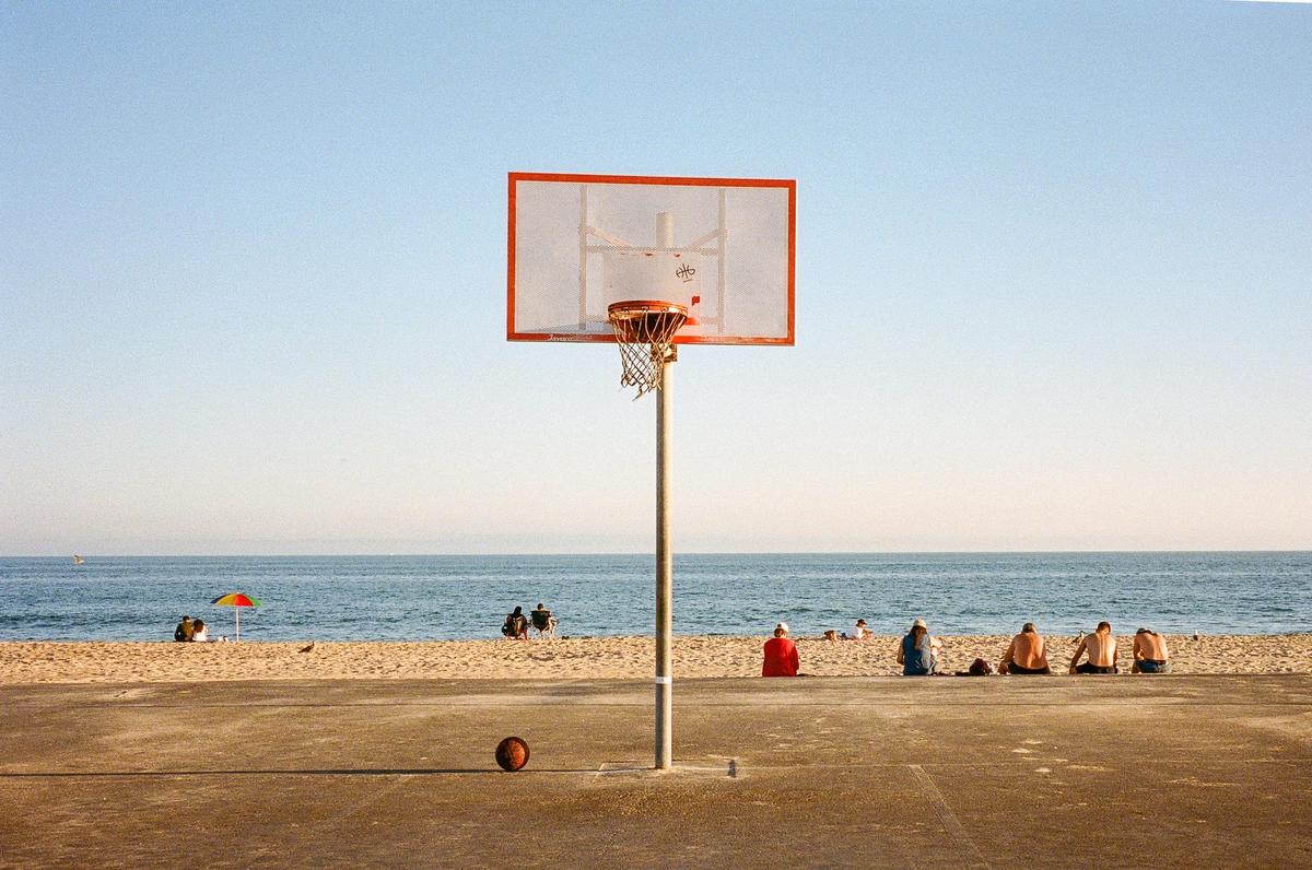 A basketball hoop stands on a court next to a beach where people are sitting by the ocean.