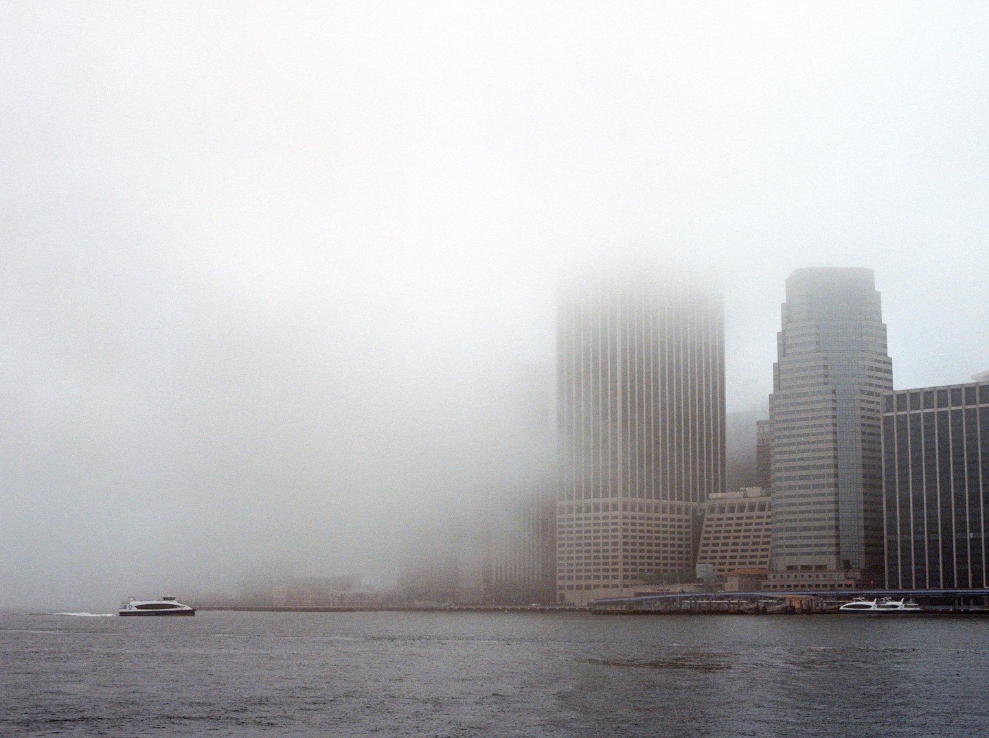 A ferry boat moves across a body of water with a foggy city skyline in the background.