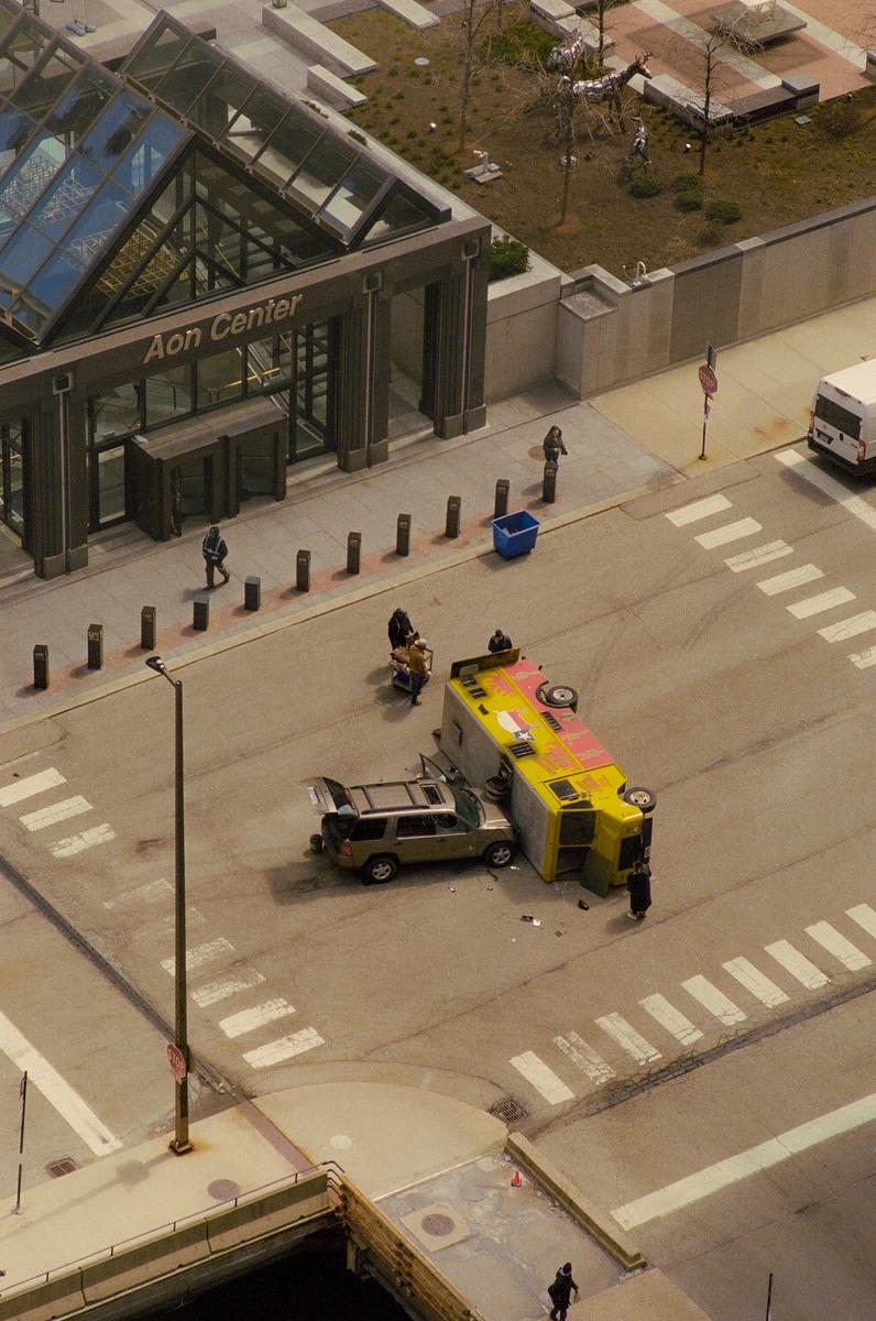 An overturned yellow truck and a car with an open trunk are on a street next to the Aon Center building.