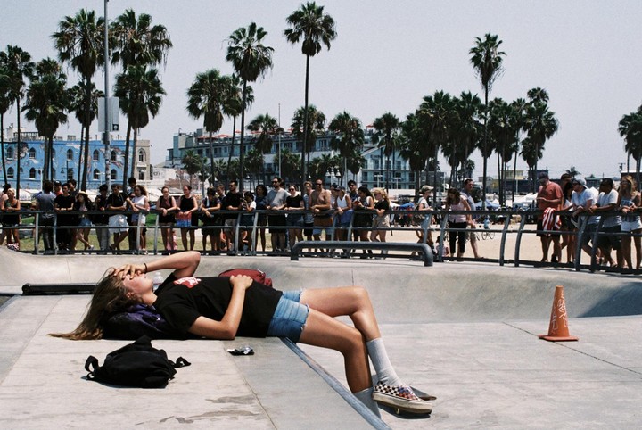 A woman lies on the edge of a skate park with palm trees and buildings in the background, while a crowd watches from behind a railing.