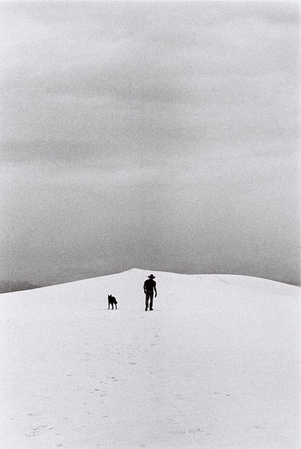 A man and a dog walk across a white sand dune under a cloudy sky.