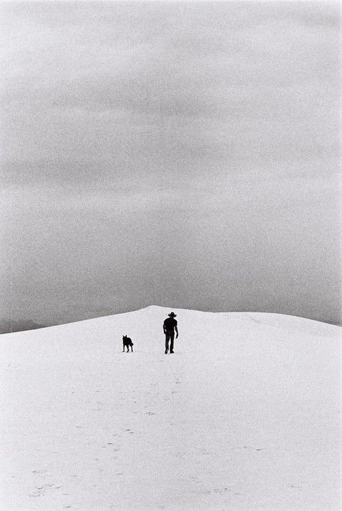 A man and a dog walk across a white sand dune under a cloudy sky.