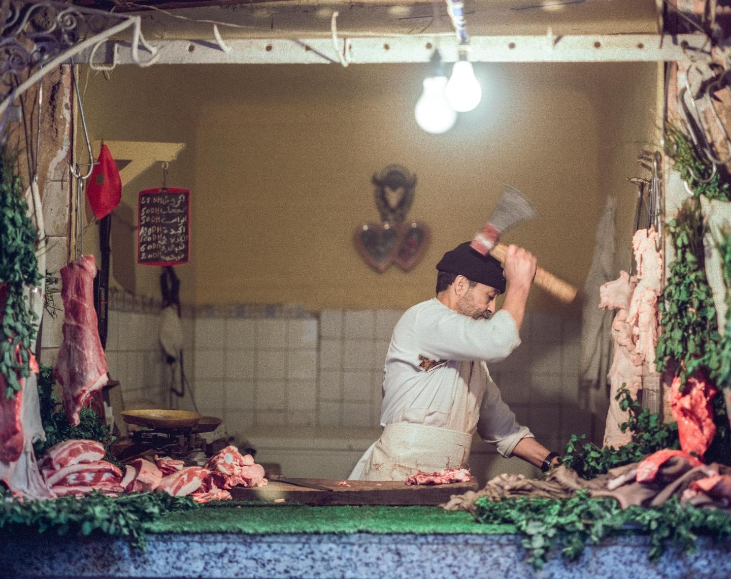 A man in a white apron and black hat chops meat with an axe in a butcher shop.