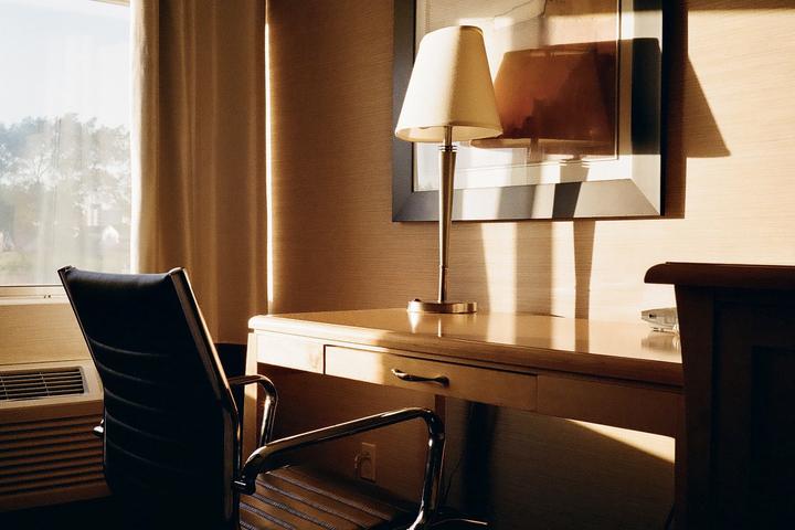 Sunlight streams through a window onto a desk with a lamp and a chair in a hotel room.