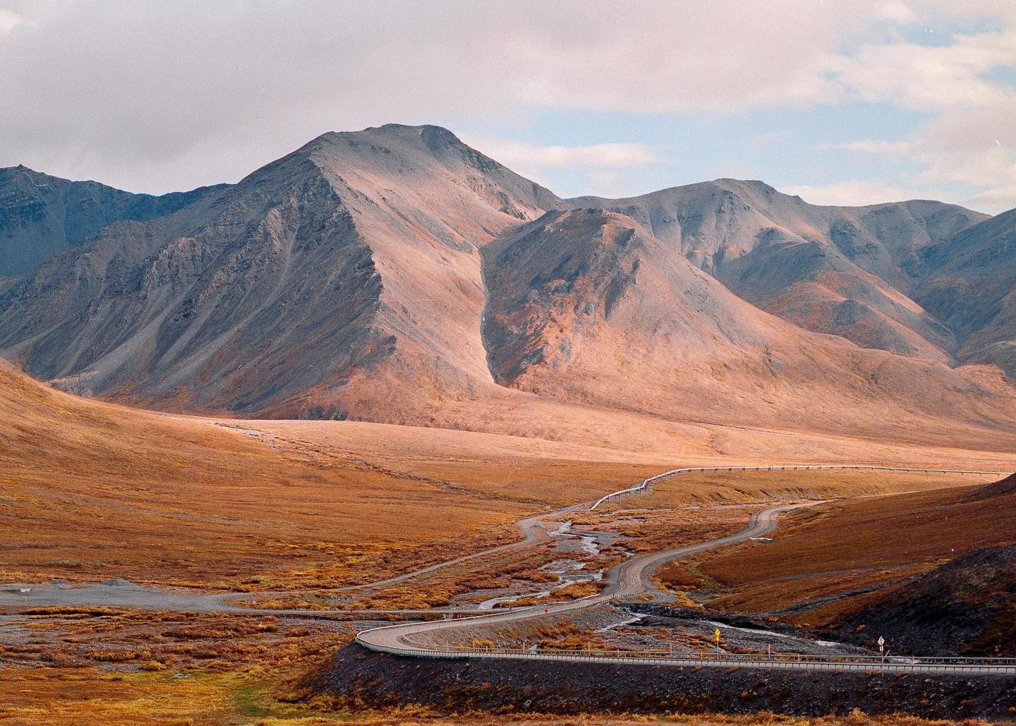 A winding road and pipeline cut through a vast, mountainous landscape with autumn foliage under a cloudy sky.