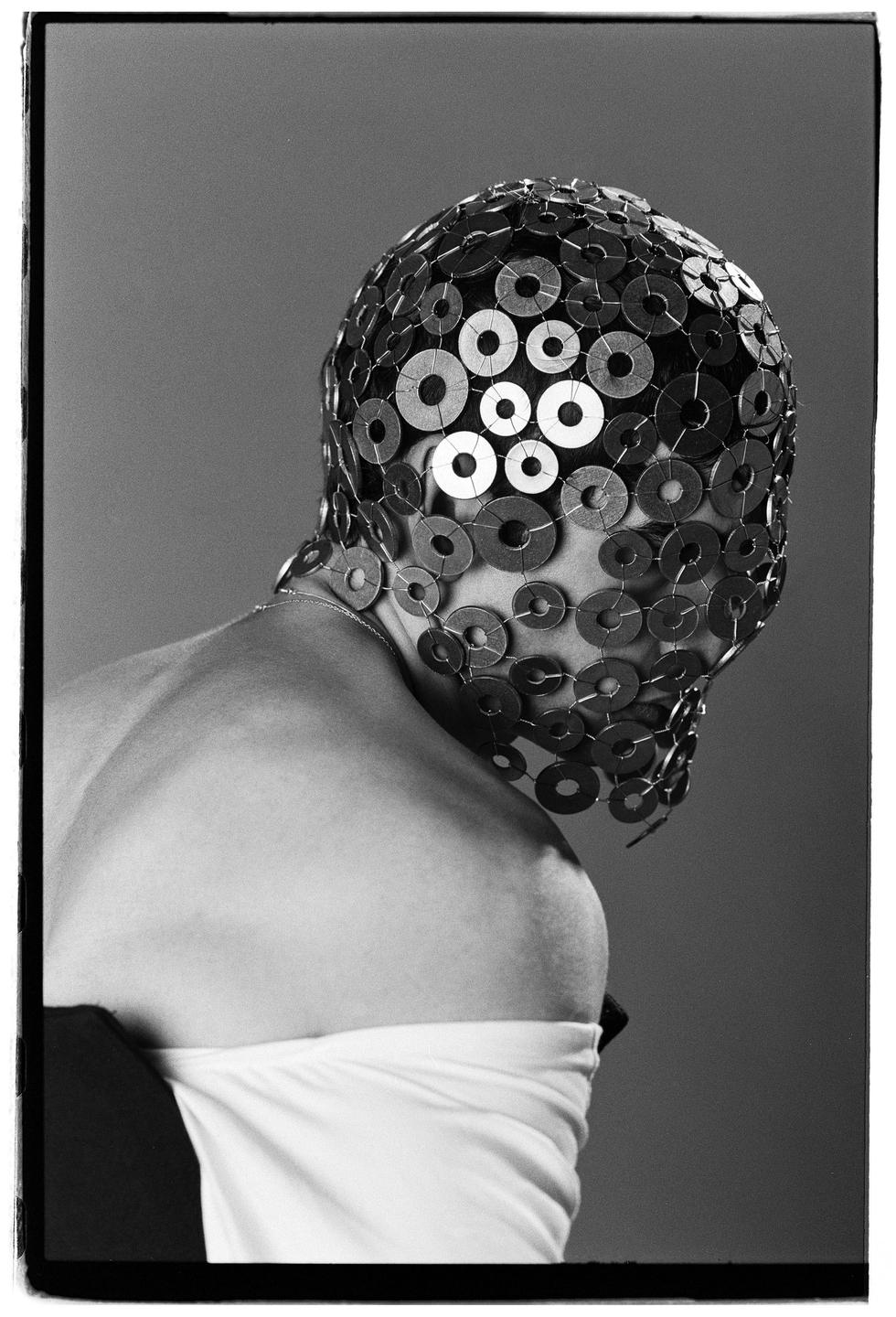A person wears a mask made of metal washers and wire, with their bare shoulder and a white garment visible.
