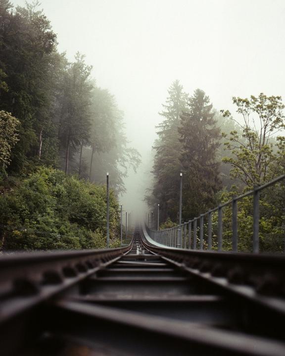 A railway track ascends a steep incline into a dense fog, flanked by a forest and a metal railing.