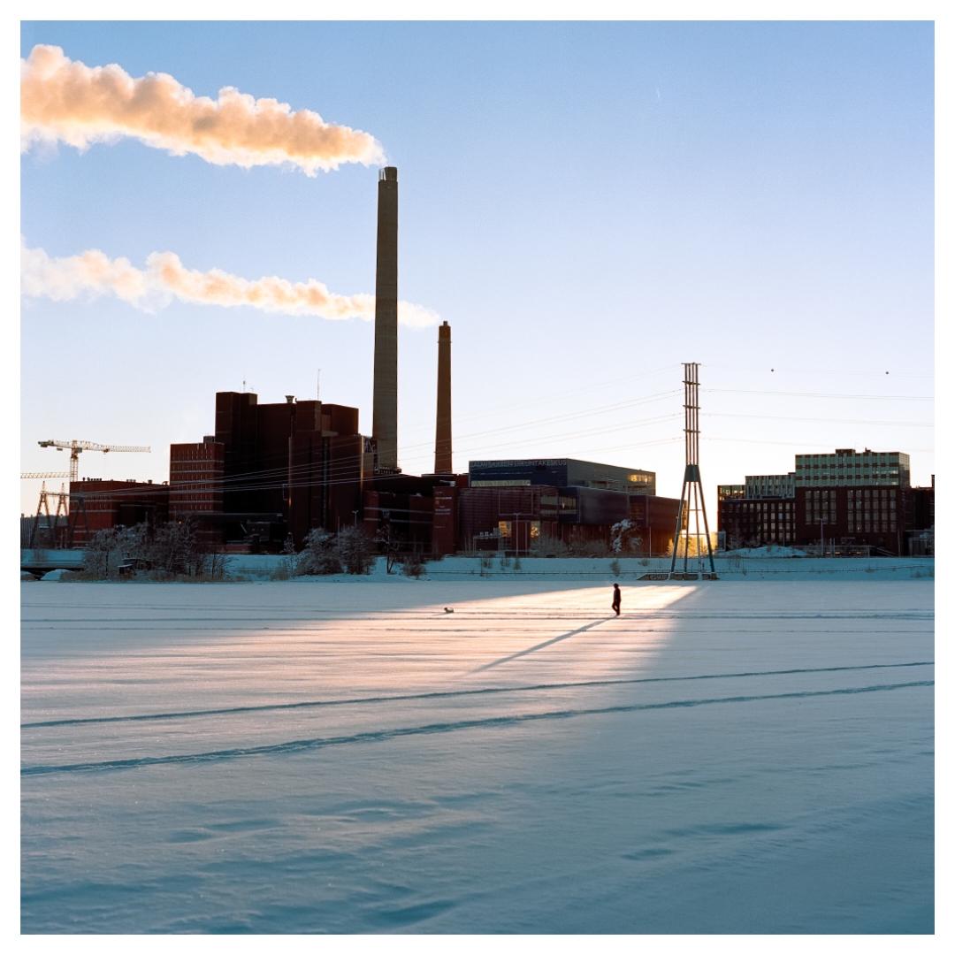 A person walks across a snow-covered field with industrial buildings and smokestacks in the background under a clear sky.