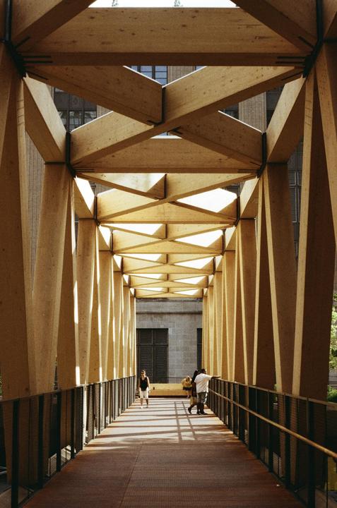 People walk along a pedestrian bridge with a wooden lattice roof that casts shadows on the walkway.
