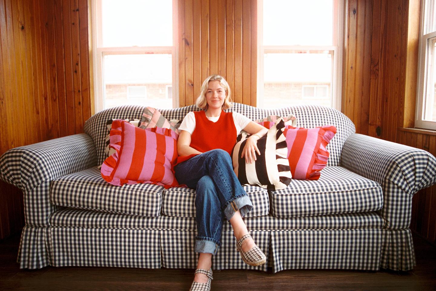 A woman sits on a gingham sofa with colorful pillows in a room with wood paneling.