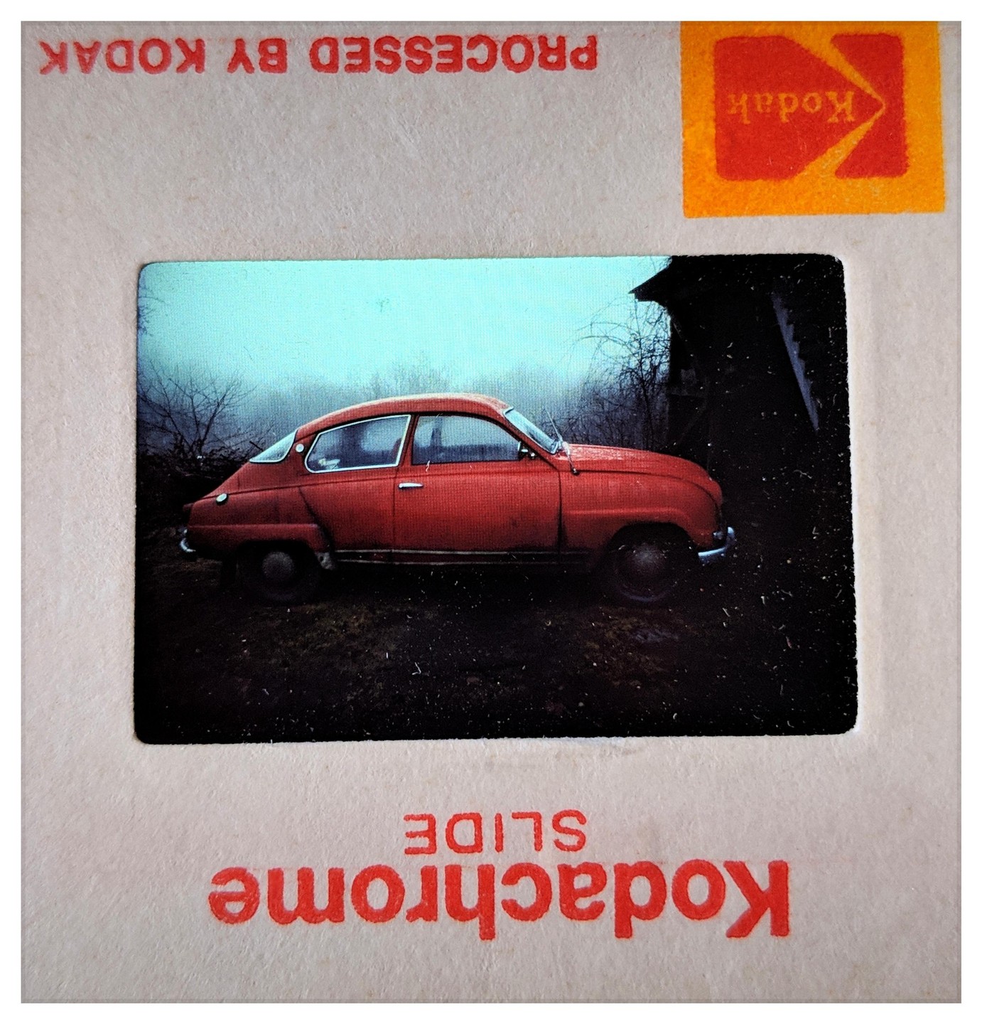 A red car is parked on a dirt road next to a building with a hazy sky in the background.