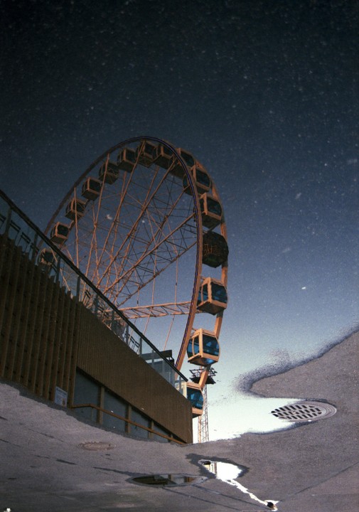 A ferris wheel is reflected in a puddle on the ground next to a building.
