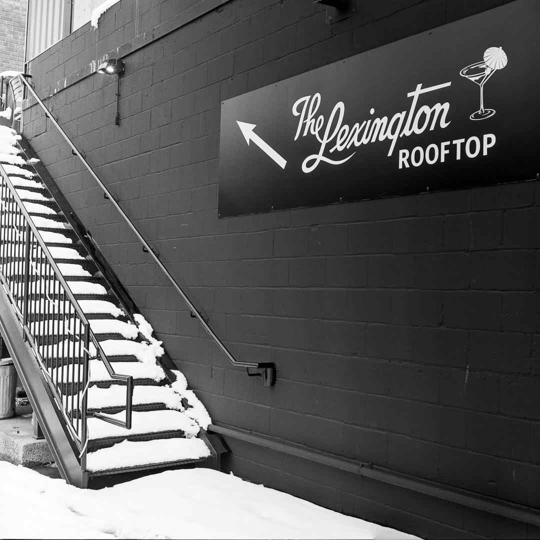 A snow-covered staircase leads up to a rooftop bar sign on a brick wall.