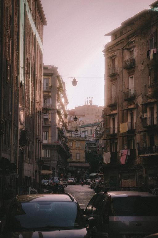 A narrow street in Naples is lined with tall, old buildings and parked cars, with laundry hanging from balconies.